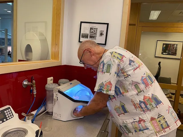 Per står ved en benk og arbeider med hendene inni en maskin som avgir blått lys i et klinisk rom