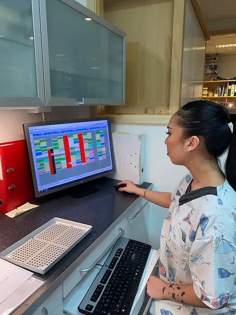 Kvinne i arbeidsklær står ved en benk og ser på en dataskjerm med fargerikt skjema på tannlegekontor