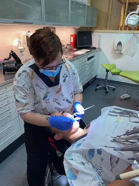 Tannlege behandler pasient i tannlegestol med munnbind og hansker, omgitt av instrumenter i klinikkrom.