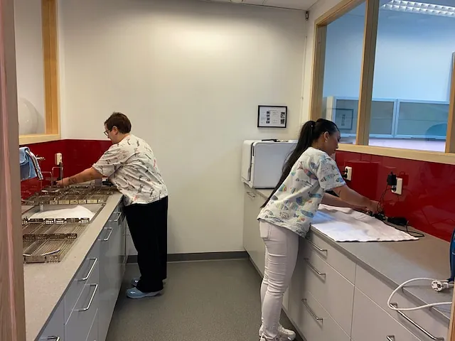 To tannleger i arbeidsklær står ved hver sin benk og rengjør utstyr i et lyst og ryddig laboratorierom.