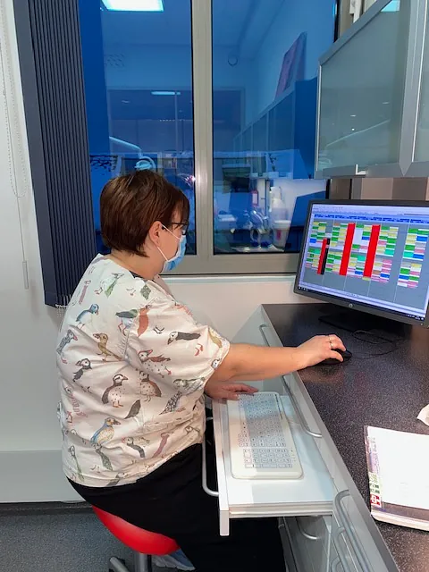 Kvinne i arbeidsklær står ved en benk og ser på en dataskjerm med fargerikt skjema på tannlegekontor